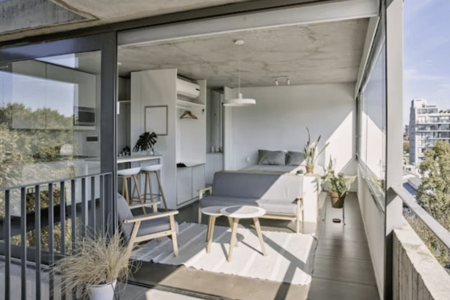 An open-plan studio apartment with a balcony and concrete ceiling on a sunny day.